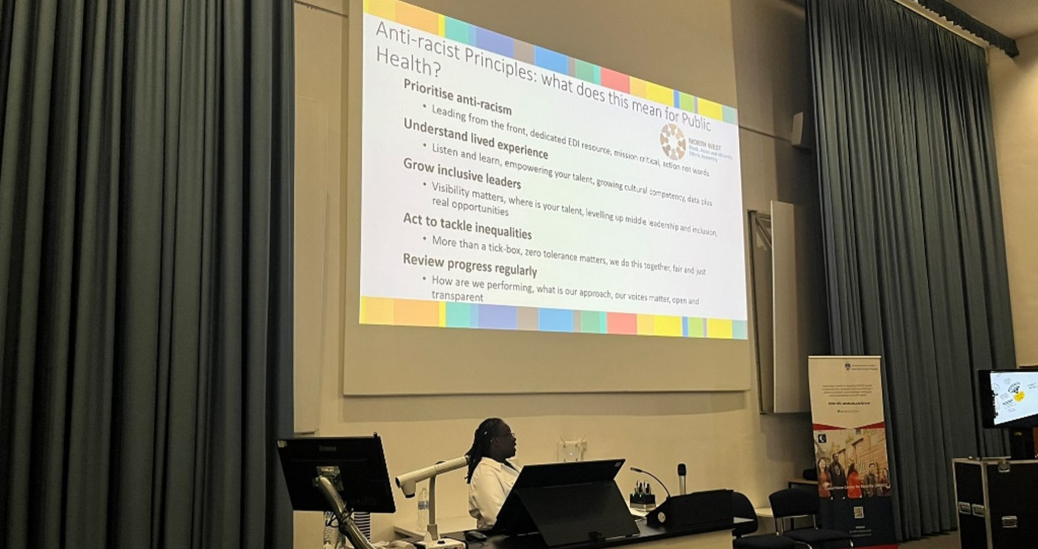 Person stands at a lecturn in front of projected screen. 