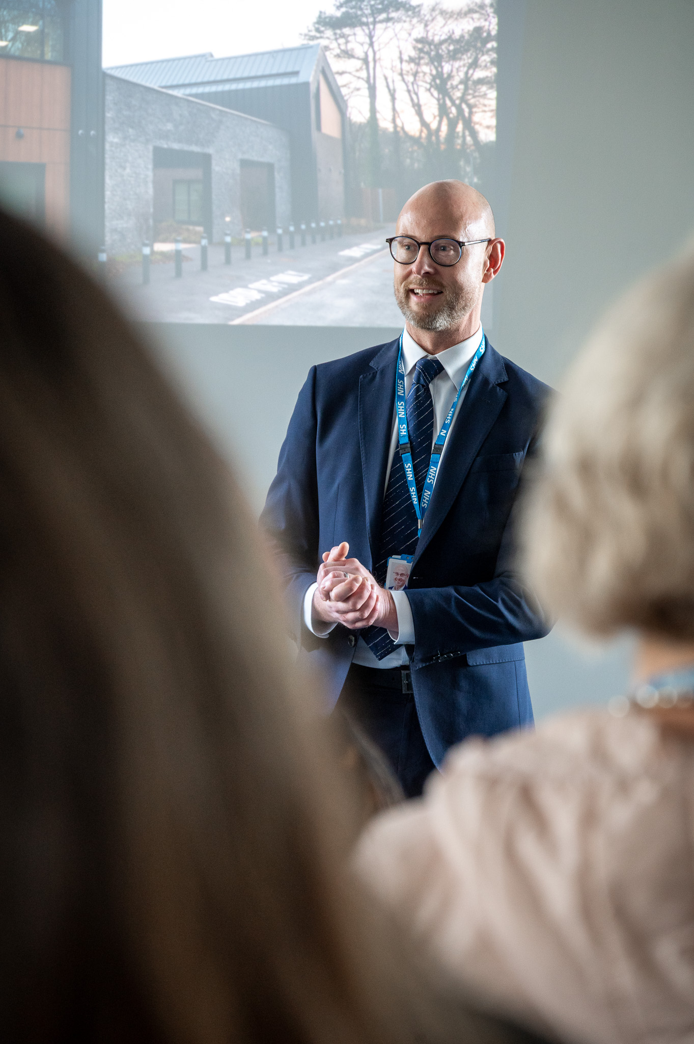 Man standing at the front of a room during an event presenting infront of a slideshow 