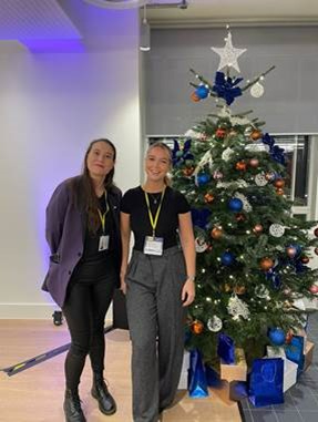 Two people stand by a Christmas tree.