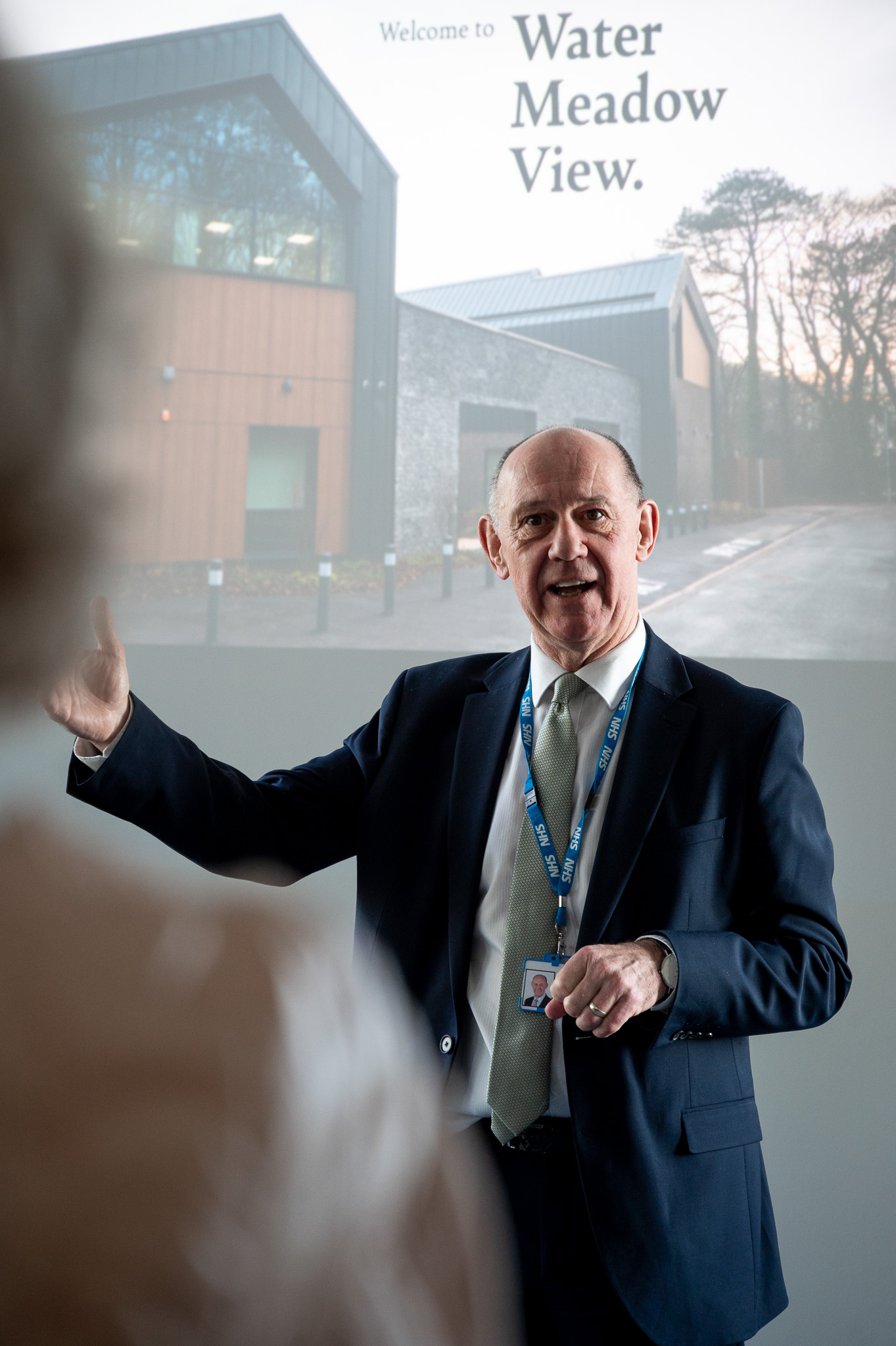 Man standing at the front of a room during an event presenting infront of a slideshow 